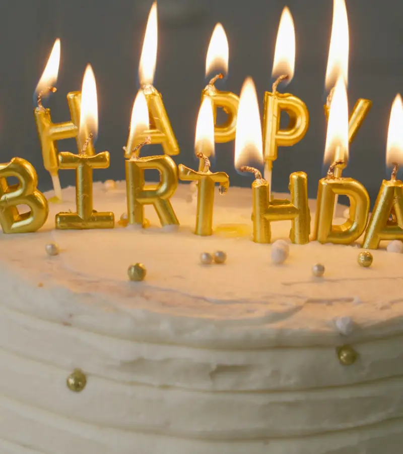 A white birthday cake with gold letter candles spelling Happy Birthday, all lit.